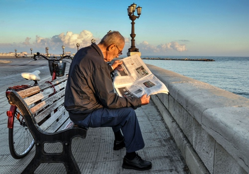 Immagine con giornale, panchina, mare, bicicletta, nuvole, lampione, occhiali, lungomare, lampioni, acqua, uomo, lettura, cielo, leggere, parapetto, persona