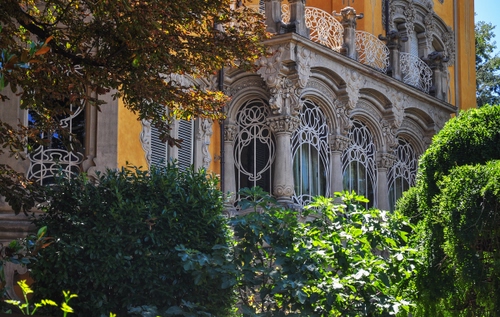 Immagine con liberty, verde, finestre, colonne, palazzo, piante, balcone, rami, foglie, ringhiera