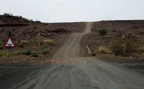 Immagine con strada, segnale, sterrata, dossi, cespugli, arbusti, pietre, cunetta, cunette