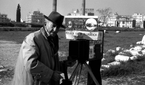 Immagine con bianconero, cappello, fotografo, anziano, uomo, cavalletto, erba, case, cartello, fotocamera, pietre, prato, cappotto, alberi, vecchio, palazzi, sassi, paese, scritta, massi