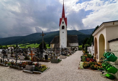 Immagine con cimitero, campanile, tombe, croci, chiesa, nuvole, lapidi, montagne, archi, fiori, annaffiatoi, verde, croce, cielo, innaffiatoi, ghiaia