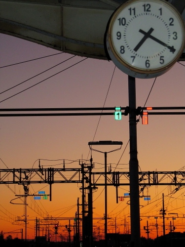 Immagine con orologio, tramonto, numeri, stazione, lancette, pali, fili, tralicci, cavi, tempo, ore, rosso, quadrante, bianco