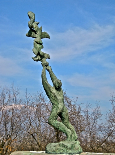 Immagine con alberi, cielo, statua, rami, nuvole, uomo, monumento, verde, scultura, uccelli, bronzo, azzurro, spogli, braccia, volo