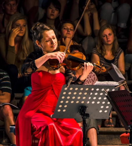 Immagine con rosso, violino, violinista, musicista, pubblico, leggio, spartito, musica, donne, archetto, mani, vestito, abito, spettatori, persone