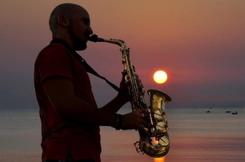 Immagine con tramonto, sassofono, sole, mare, musicista, uomo, sassofonista, strumento, barche, musica, ottone, controluce, acqua, giallo, sax, profilo