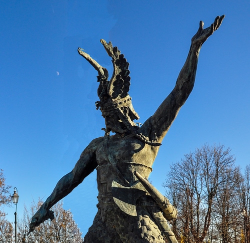 Immagine con statua, alberi, cielo, elmo, braccia, guerriero, ali, lampione, monumento, ascia, bronzo, azzurro, scultura, corona, rami